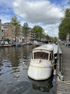A picturesque canal scene features a small white boat moored to a wooden dock. The boat has a 'Tourist' sign and decorative elements. The water reflects the surroundings, including rows of traditional brick buildings with large windows. Trees line the canal, and a partly cloudy sky adds to the ambiance.