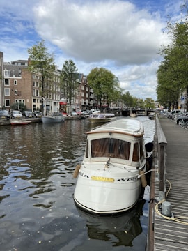 A picturesque canal scene features a small white boat moored to a wooden dock. The boat has a 'Tourist' sign and decorative elements. The water reflects the surroundings, including rows of traditional brick buildings with large windows. Trees line the canal, and a partly cloudy sky adds to the ambiance.