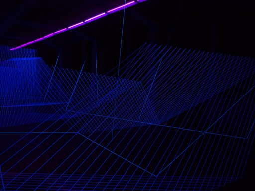 Multiple intersecting blue luminescent lines form a geometric mesh structure on a dark background. A purple neon light strip runs horizontally across the top portion of the image, casting a glow.