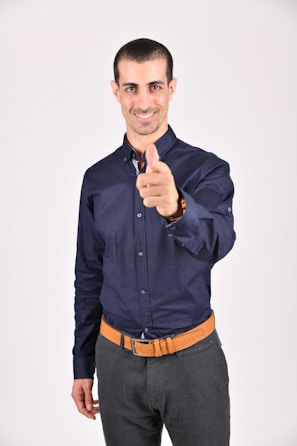 A man with short dark hair and a friendly smile is pointing toward the camera. He is wearing a dark blue button-up shirt, dark pants, and a tan belt. The background is plain and light-colored.