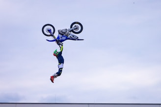 A motorbike rider is performing a daring stunt, hanging upside down in mid-air while gripping the handlebar of a motorcycle with one hand. The motorcycle is also inverted, indicating a high-flying acrobatic trick. The sky in the background is overcast, emphasizing the focus on the stunt.