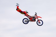 A person is performing a stunt on a motocross bike mid-air. They are wearing bright red and yellow protective gear, and the bike is primarily red with blue rims. The background is a clear sky.