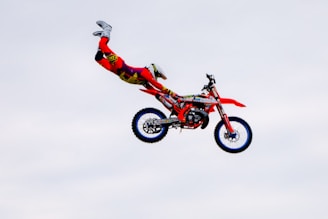 A person is performing a stunt on a motocross bike mid-air. They are wearing bright red and yellow protective gear, and the bike is primarily red with blue rims. The background is a clear sky.