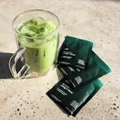 A clear glass mug filled with iced matcha latte sits on a textured, light-colored surface. Three green packaging sachets labeled 'energy + focus' lie nearby, slightly overlapping each other.