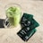 A clear glass mug filled with iced matcha latte sits on a textured, light-colored surface. Three green packaging sachets labeled 'energy + focus' lie nearby, slightly overlapping each other.
