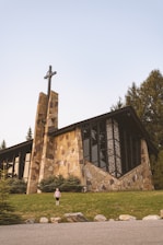 A stone church with large windows and a cross on top sits amidst a well-maintained grassy area. Evergreen trees surround the church, and a young child stands on the grass in front.