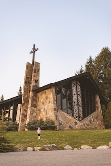 A stone church with large windows and a cross on top sits amidst a well-maintained grassy area. Evergreen trees surround the church, and a young child stands on the grass in front.