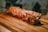 A large skewer of roasted meat rests on a wooden board. The meat appears to be perfectly cooked, displaying a range of golden brown and crispy textures, with visible grill marks. The background is softly blurred, emphasizing the focus on the skewered meat.