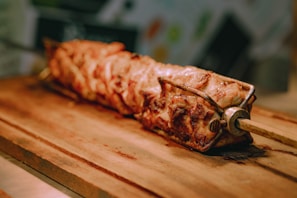 A large skewer of roasted meat rests on a wooden board. The meat appears to be perfectly cooked, displaying a range of golden brown and crispy textures, with visible grill marks. The background is softly blurred, emphasizing the focus on the skewered meat.