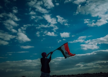 A person raises an Indian flag against a backdrop of a vast blue sky filled with scattered white clouds. The silhouette of the person adds a dramatic effect to the scene.