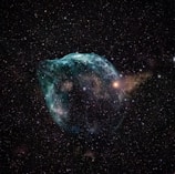 A captivating image of a nebula set against the vast expanse of space. The nebula features swirling clouds of gas and dust, primarily in hues of blue and green, with some reddish and yellowish regions. Numerous stars of varying brightness are scattered across the background.
