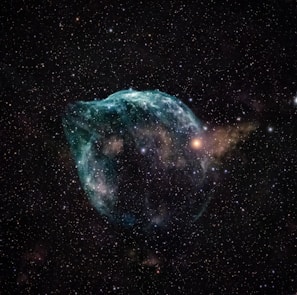 A captivating image of a nebula set against the vast expanse of space. The nebula features swirling clouds of gas and dust, primarily in hues of blue and green, with some reddish and yellowish regions. Numerous stars of varying brightness are scattered across the background.