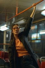 A person wearing sunglasses and stylish clothing is standing inside a dimly lit bus. They are holding onto an overhead rail with one hand, exuding a sense of confidence and fashion-forward attitude. The person is dressed in a dark coat over an orange shirt, and the bus interior features red seats and metallic handrails.
