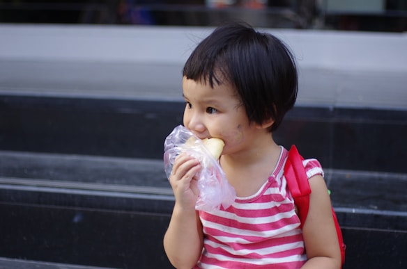 A young child with short dark hair is holding a plastic-wrapped snack, possibly bread, and is taking a bite. The child is wearing a pink and white striped sleeveless shirt and a red backpack. They are sitting on dark gray steps, looking to the side.