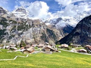 A picturesque village nestled in a valley among towering snow-capped mountains. Traditional wooden houses are scattered across lush green fields dotted with yellow flowers. A winding path leads through the village, which is surrounded by dense forest at the base of steep rocky cliffs.