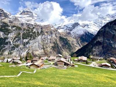 A picturesque village nestled in a valley among towering snow-capped mountains. Traditional wooden houses are scattered across lush green fields dotted with yellow flowers. A winding path leads through the village, which is surrounded by dense forest at the base of steep rocky cliffs.