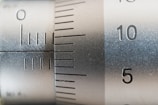 A close-up view of a metallic measuring instrument, possibly a micrometer or caliper, featuring engraved numerical markings and increment lines. The focus is on numbers 5 and 10, with a brushed metal texture visible.