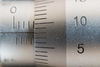 A close-up view of a metallic measuring instrument, possibly a micrometer or caliper, featuring engraved numerical markings and increment lines. The focus is on numbers 5 and 10, with a brushed metal texture visible.