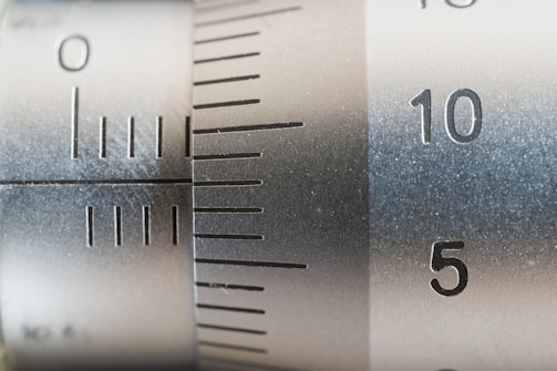 A close-up view of a metallic measuring instrument, possibly a micrometer or caliper, featuring engraved numerical markings and increment lines. The focus is on numbers 5 and 10, with a brushed metal texture visible.