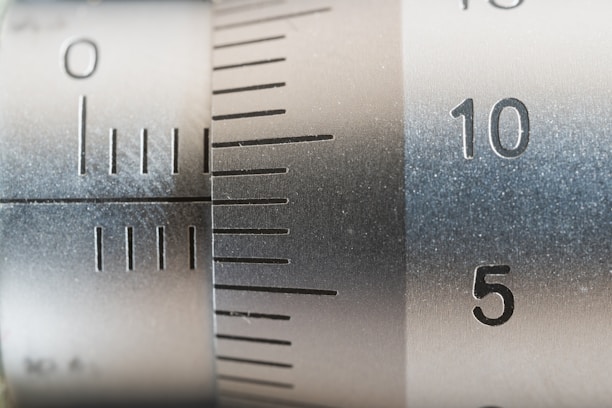 A close-up view of a metallic measuring instrument, possibly a micrometer or caliper, featuring engraved numerical markings and increment lines. The focus is on numbers 5 and 10, with a brushed metal texture visible.
