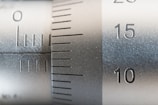 Close-up of a metallic measuring instrument with engraved numbers and lines, possibly a caliper or micrometer. The numbers 10, 15, and 20 are clearly visible along with measurement lines, indicating precision engineering.