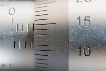 Close-up of a metallic measuring instrument with engraved numbers and lines, possibly a caliper or micrometer. The numbers 10, 15, and 20 are clearly visible along with measurement lines, indicating precision engineering.