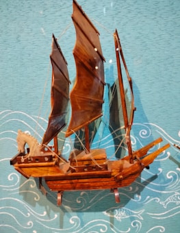 A wooden ship model is displayed against a blue textured backdrop decorated with white wave and fish designs. The model features intricately crafted brown sails and a carved wooden horse figure at the front.