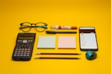 A collection of office and school supplies is neatly arranged on a bright yellow background. The items include a pair of eyeglasses, a calculator, two blocks of sticky notes in pastel colors, a smartphone displaying a list, two pencils, a pen, a white and red correction pen, and a small pencil sharpener with a colorful design.