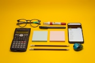 A collection of office and school supplies is neatly arranged on a bright yellow background. The items include a pair of eyeglasses, a calculator, two blocks of sticky notes in pastel colors, a smartphone displaying a list, two pencils, a pen, a white and red correction pen, and a small pencil sharpener with a colorful design.