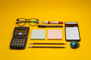 A collection of office and school supplies is neatly arranged on a bright yellow background. The items include a pair of eyeglasses, a calculator, two blocks of sticky notes in pastel colors, a smartphone displaying a list, two pencils, a pen, a white and red correction pen, and a small pencil sharpener with a colorful design.