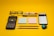 A collection of office and school supplies is neatly arranged on a bright yellow background. The items include a pair of eyeglasses, a calculator, two blocks of sticky notes in pastel colors, a smartphone displaying a list, two pencils, a pen, a white and red correction pen, and a small pencil sharpener with a colorful design.
