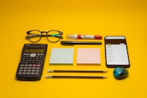 A collection of office and school supplies is neatly arranged on a bright yellow background. The items include a pair of eyeglasses, a calculator, two blocks of sticky notes in pastel colors, a smartphone displaying a list, two pencils, a pen, a white and red correction pen, and a small pencil sharpener with a colorful design.