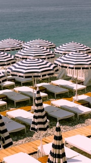 A beachfront scene with neatly arranged rows of striped sun umbrellas and white sunbeds on a pebbled beach. The calm, blue ocean is visible in the background, creating a serene and organized setting.