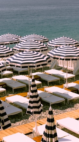A beachfront scene with neatly arranged rows of striped sun umbrellas and white sunbeds on a pebbled beach. The calm, blue ocean is visible in the background, creating a serene and organized setting.