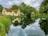 A quaint cottage with a thatched roof sits beside a calm, reflective body of water surrounded by lush green vegetation and trees. The serene scene is under a partly cloudy sky, with the cottage and trees mirrored in the still water.