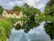 A quaint cottage with a thatched roof sits beside a calm, reflective body of water surrounded by lush green vegetation and trees. The serene scene is under a partly cloudy sky, with the cottage and trees mirrored in the still water.