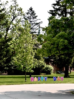 A lush green park features tall trees and scattered shrubs, with colorful letters planted on the grass spelling out the phrase 'HAPPY DAY'. The scene is bright and sunny, suggesting a pleasant outdoor atmosphere.