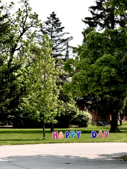 A lush green park features tall trees and scattered shrubs, with colorful letters planted on the grass spelling out the phrase 'HAPPY DAY'. The scene is bright and sunny, suggesting a pleasant outdoor atmosphere.