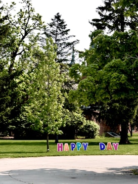 A lush green park features tall trees and scattered shrubs, with colorful letters planted on the grass spelling out the phrase 'HAPPY DAY'. The scene is bright and sunny, suggesting a pleasant outdoor atmosphere.