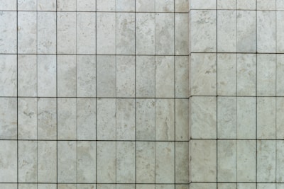 A close-up view of a tiled wall featuring rectangular stone tiles arranged in a grid pattern with visible grout lines. The surface texture includes subtle variations and natural speckles typical of stone.