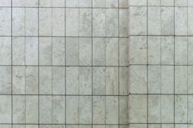 A close-up view of a tiled wall featuring rectangular stone tiles arranged in a grid pattern with visible grout lines. The surface texture includes subtle variations and natural speckles typical of stone.