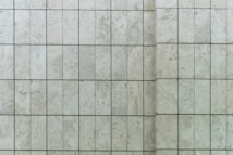 A close-up view of a tiled wall featuring rectangular stone tiles arranged in a grid pattern with visible grout lines. The surface texture includes subtle variations and natural speckles typical of stone.