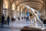A grand gallery with a high, arched ceiling and large sculptures displayed along the sides on pedestals. There is a prominent classical marble statue in the foreground, depicting a dynamic figure in motion. Several people are walking and observing the artwork, adding a sense of scale and vitality to the scene.