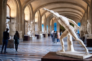 A grand gallery with a high, arched ceiling and large sculptures displayed along the sides on pedestals. There is a prominent classical marble statue in the foreground, depicting a dynamic figure in motion. Several people are walking and observing the artwork, adding a sense of scale and vitality to the scene.