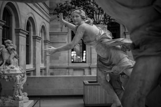 A classical sculpture of a dynamic, flowing figure is set within an elegant indoor setting. The figure exudes movement, draped in detailed robes and captured in mid-motion. The setting includes ornate architectural elements such as arches and windows, with another less detailed sculpture visible nearby.