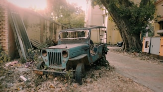 A battered, old jeep is parked in an overgrown and neglected area. The vehicle is rusted, missing its windshield, and surrounded by fallen leaves and rubble. It is placed beside a large tree and a dilapidated brick wall, with a narrow pathway leading past some buildings in the background. Sunlight filters through the trees, casting a warm hue over the scene.