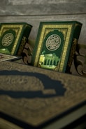 A close-up view of ornate books with intricate Arabic calligraphy and a picture of a mosque on their covers. The book covers feature a rich green background with gold embellishments.