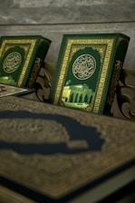 A close-up view of ornate books with intricate Arabic calligraphy and a picture of a mosque on their covers. The book covers feature a rich green background with gold embellishments.