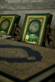 A close-up view of ornate books with intricate Arabic calligraphy and a picture of a mosque on their covers. The book covers feature a rich green background with gold embellishments.