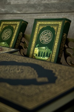 A close-up view of ornate books with intricate Arabic calligraphy and a picture of a mosque on their covers. The book covers feature a rich green background with gold embellishments.
